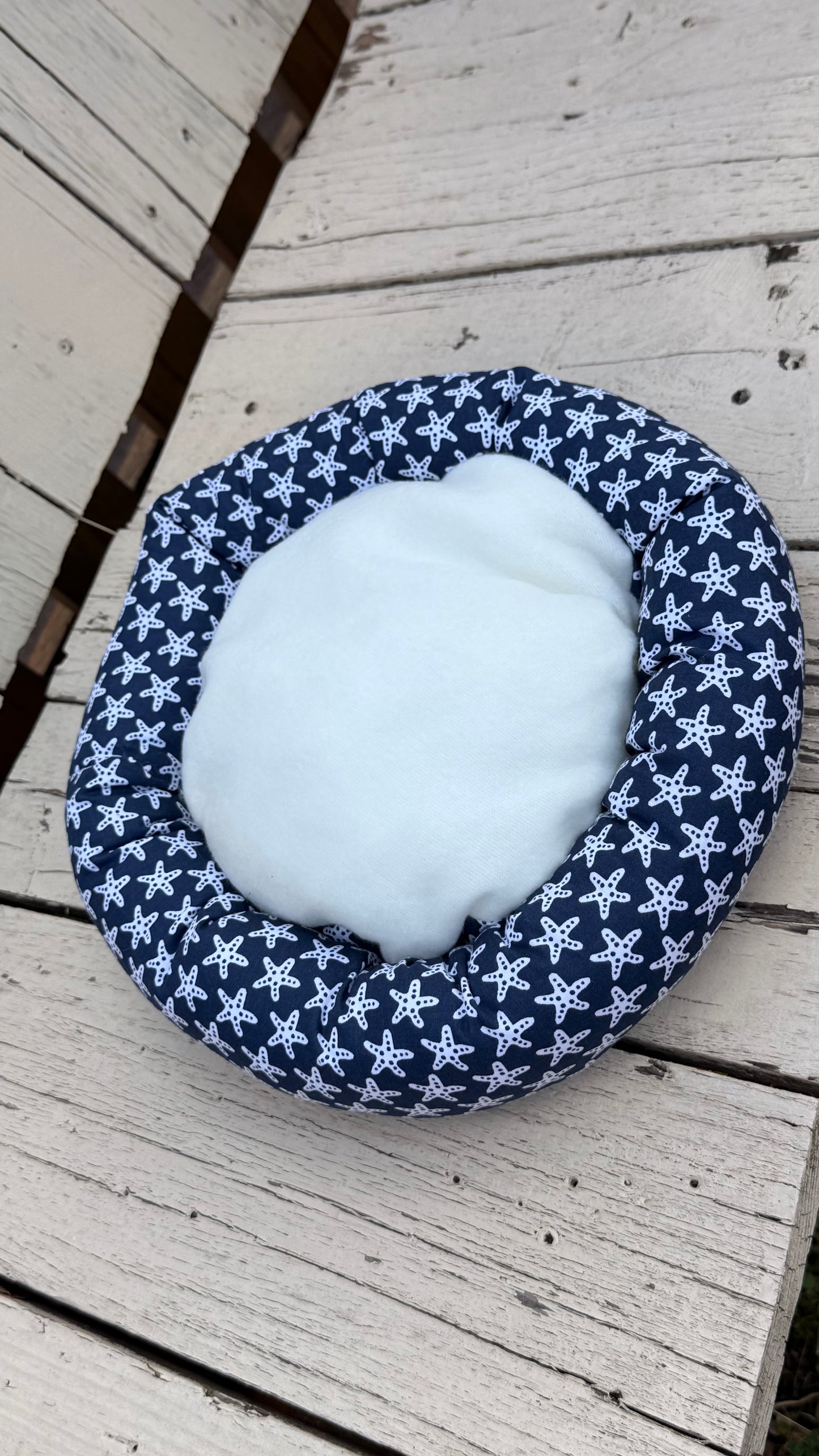 Ein runder, handgemachter Kuschel-Donut für Meerschweinchen aus dunkelblauem Baumwollstoff. Das Material ist durchgehend mit einem dichten Muster aus vielen kleinen, weißen Seesternen bedruckt. Das Kleintierbett liegt flach auf einer weiß gestrichenen Holzbank.