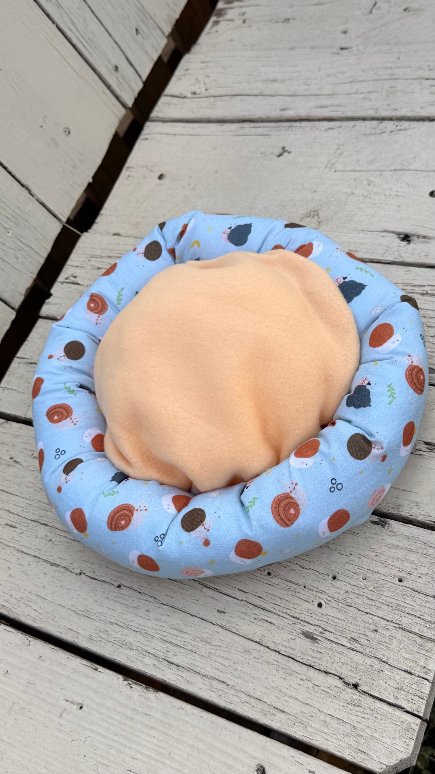 Ein runder, handgemachter Kuschel-Donut für Meerschweinchen aus hellblauem Baumwollstoff. Der Stoff ist komplett mit niedlichen Motiven von kleinen Igeln, bunten Schneckenhäusern und Pilzen bedruckt. Das gemütliche Kleintierbett liegt flach auf einer weiß gestrichenen Holzbank