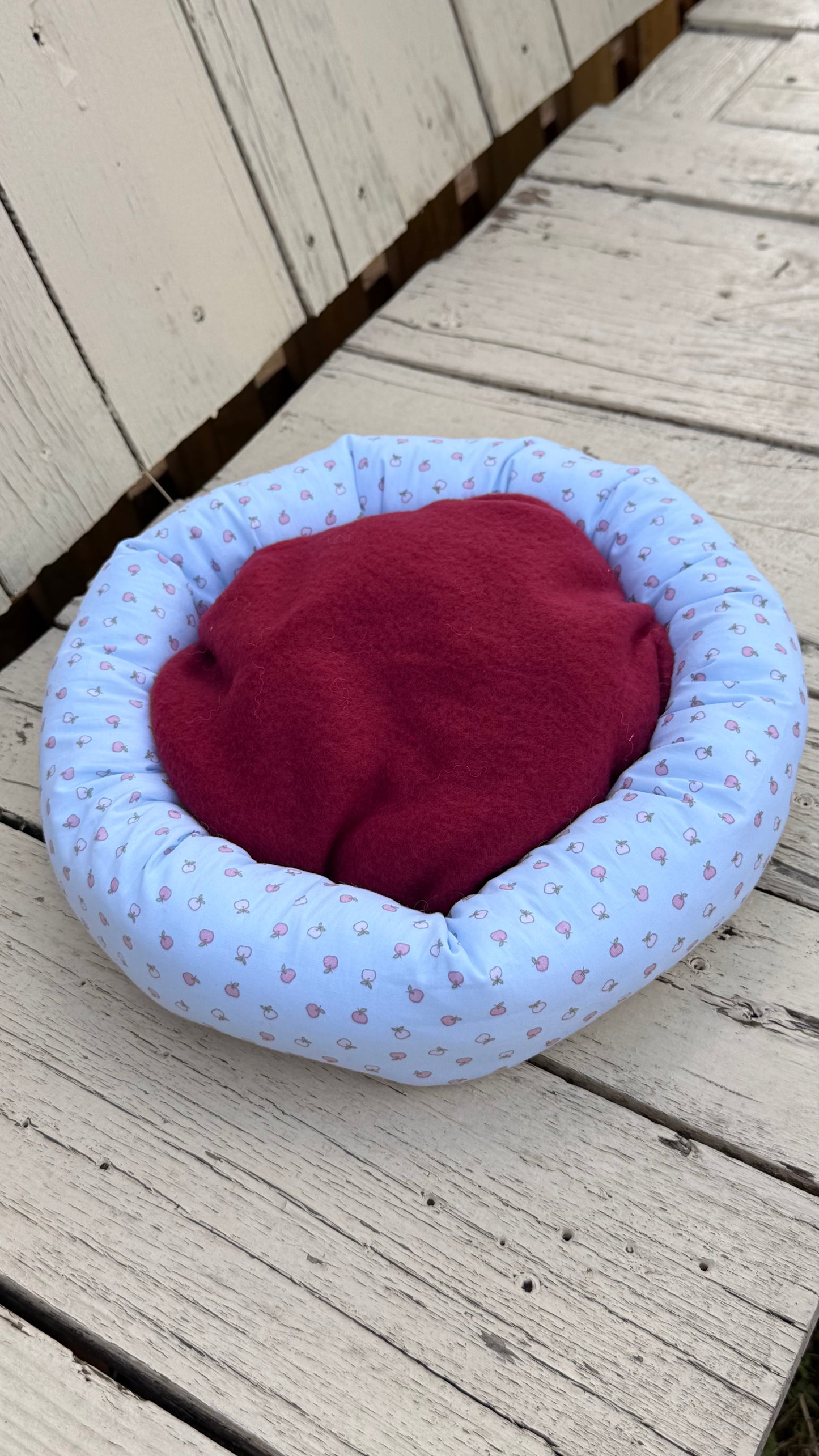 Ein hellblauer, runder Kuschel-Donut für Meerschweinchen mit einem feinen, Allover-Muster aus kleinen, rosa Äpfeln. Das handgemachte Kleintierbett liegt flach auf einer rustikalen, weiß gestrichenen Holzbank aus Holzbrettern.