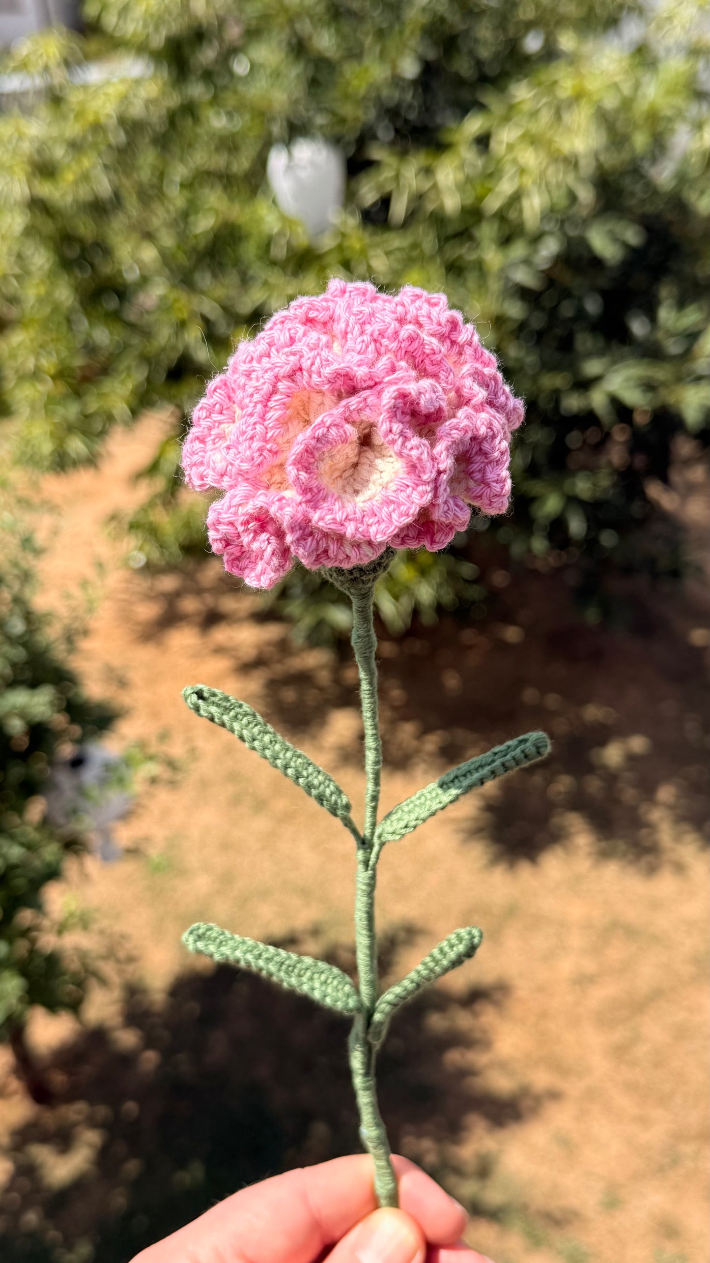Handgehäkelte Nelke in zartem Rosa und Weiß mit mehrschichtigen Blütenblättern, grünem Stiel und mehreren Blättern, handgemachte Häkelblume aus Dein Krämerladen.