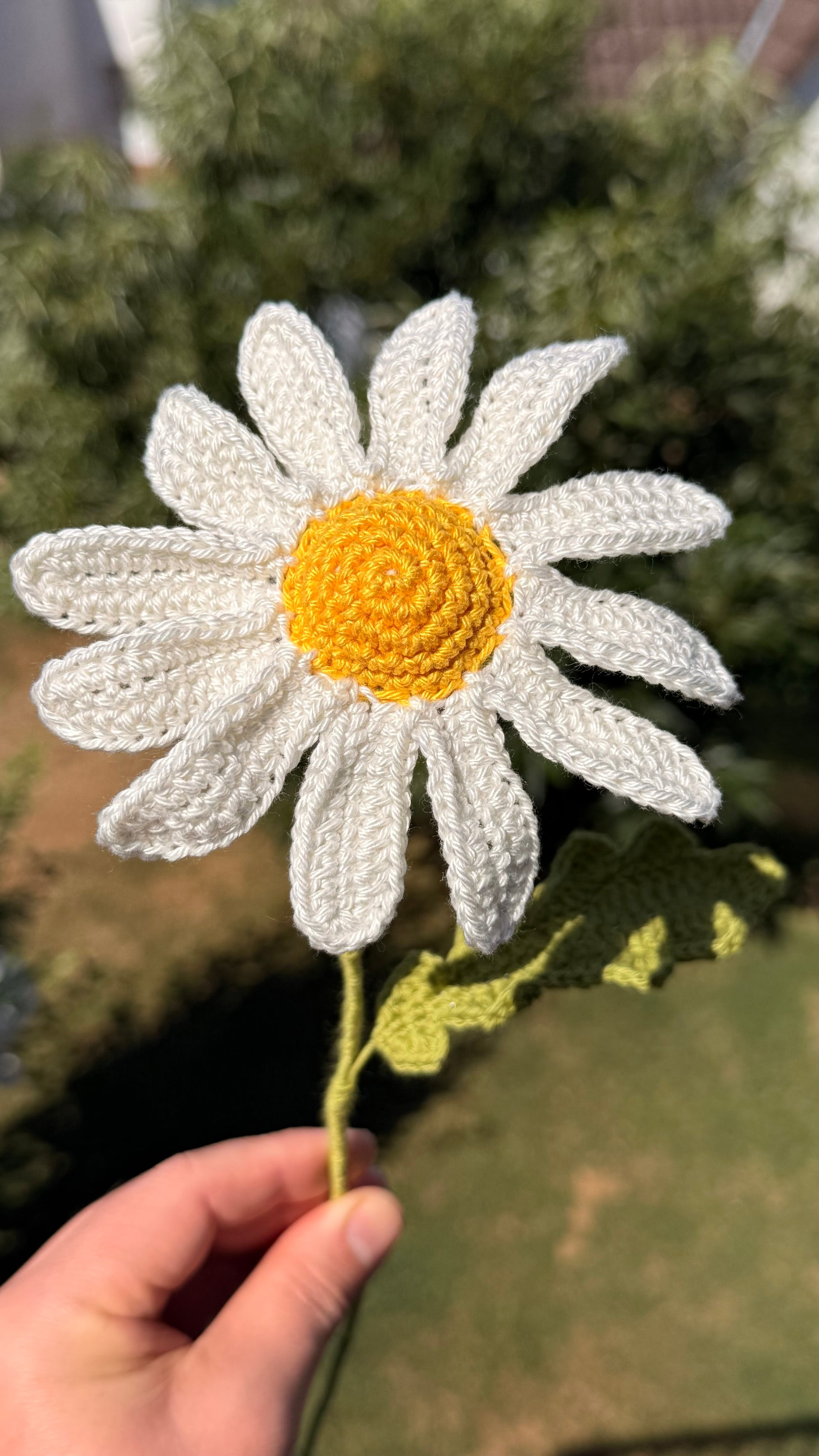 Handgehäkelte Margerite als dekorative Kunstblume, weiße Blütenblätter mit gelbem Kern und grünem Stiel, liebevolle Handarbeit für eine nachhaltige Blumendekoration.