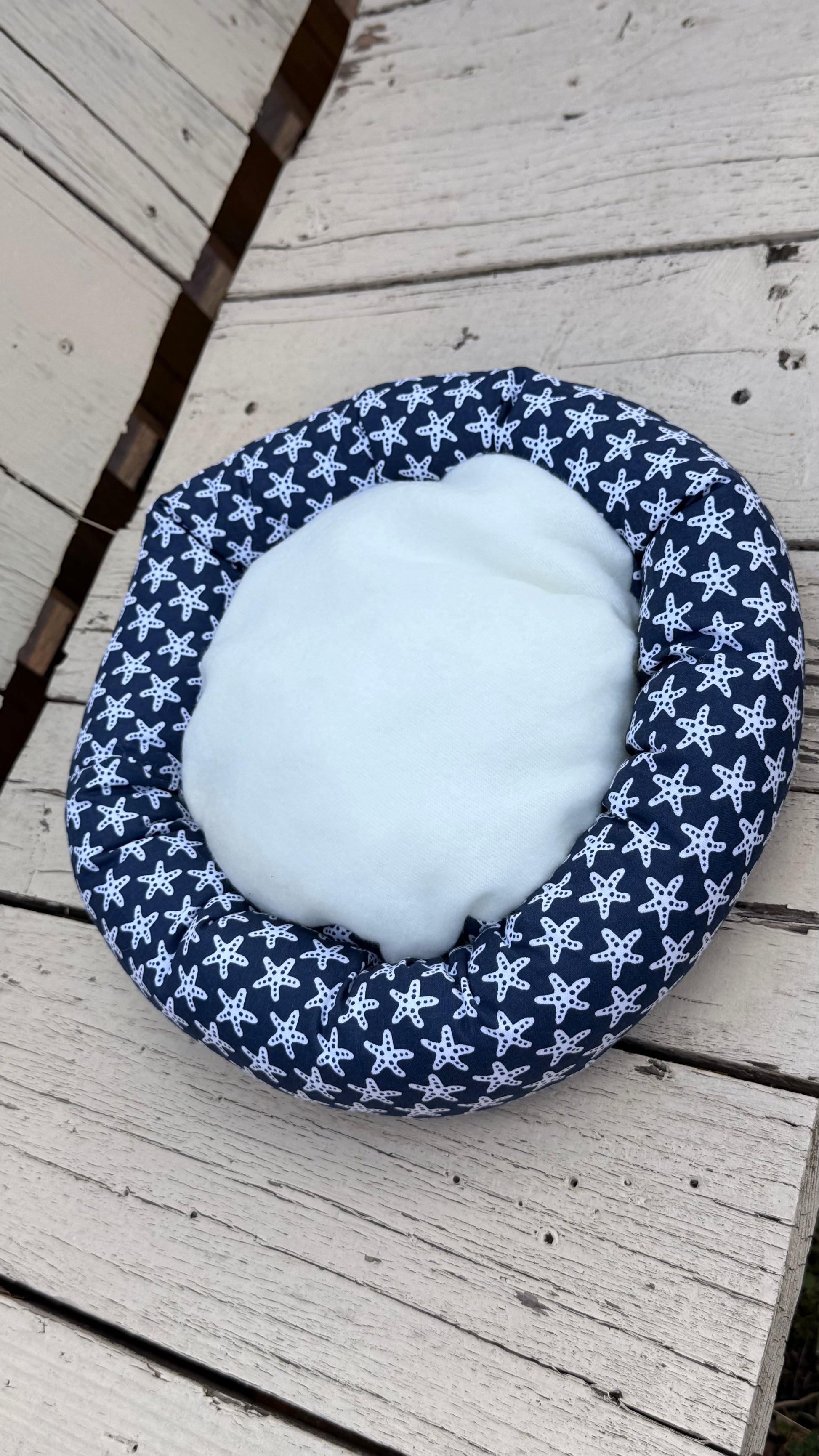 Ein runder, handgemachter Kuschel-Donut für Meerschweinchen aus dunkelblauem Baumwollstoff. Das Material ist durchgehend mit einem dichten Muster aus vielen kleinen, weißen Seesternen bedruckt. Das Kleintierbett liegt flach auf einer weiß gestrichenen Holzbank.