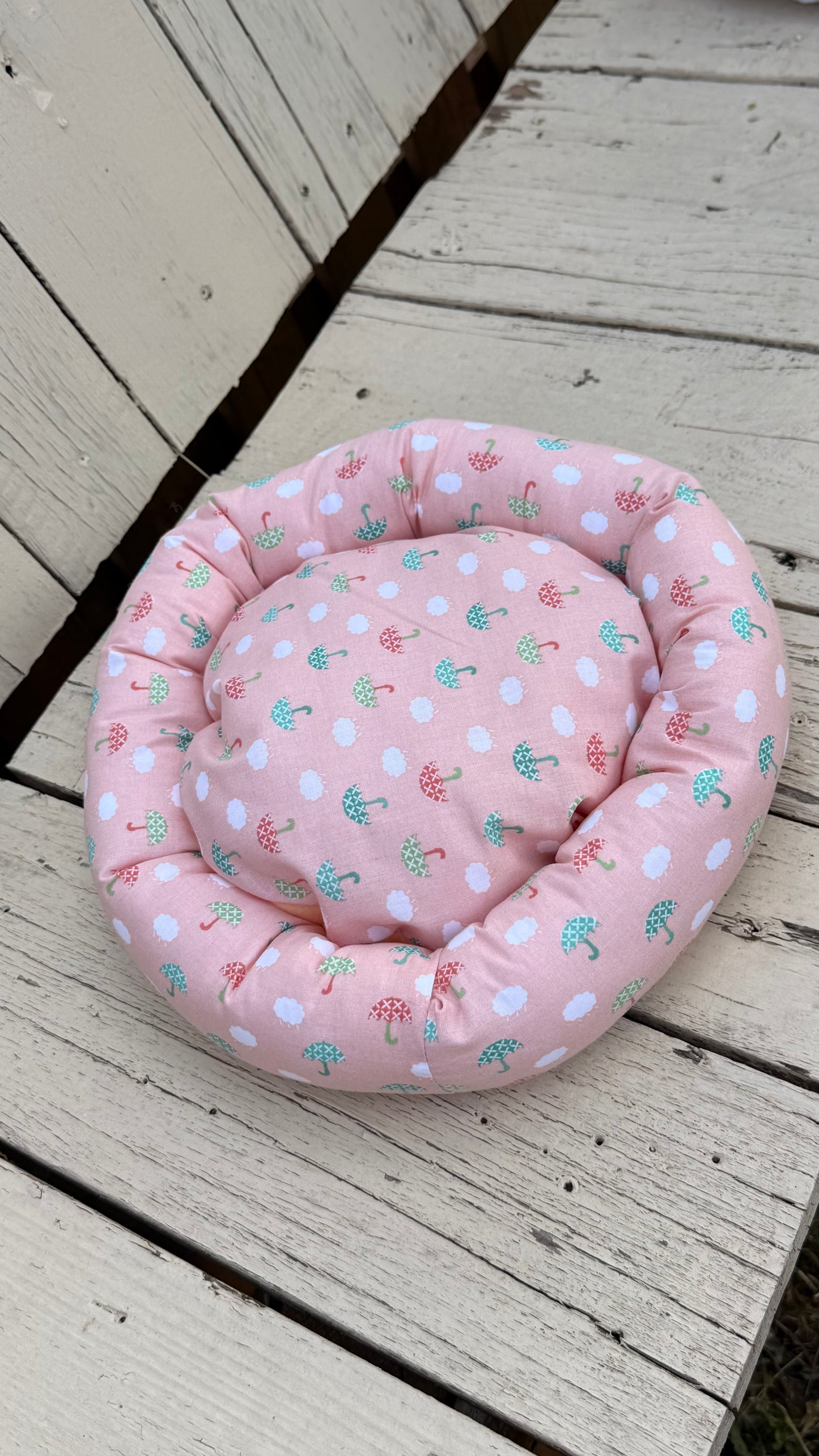 Ein runder, handgemachter Kuschel-Donut für Meerschweinchen aus altrosa Baumwollstoff. Das Material ist durchgehend mit einem Muster aus kleinen, weißen Wolken sowie grünen und roten Regenschirmen bedruckt. Das Kleintierbett liegt flach auf einer weiß gestrichenen Holzbank.