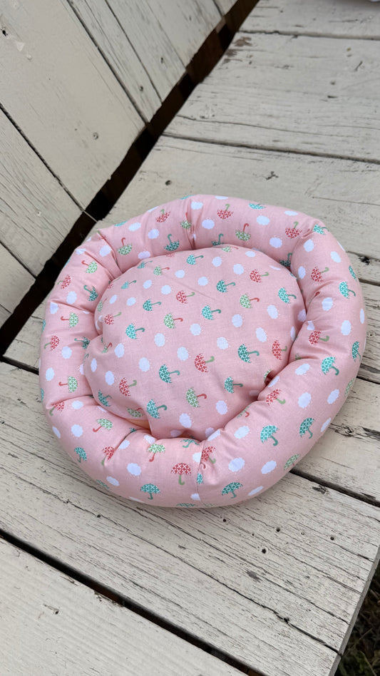 Ein runder, handgemachter Kuschel-Donut für Meerschweinchen aus altrosa Baumwollstoff. Das Material ist durchgehend mit einem Muster aus kleinen, weißen Wolken sowie grünen und roten Regenschirmen bedruckt. Das Kleintierbett liegt flach auf einer weiß gestrichenen Holzbank.