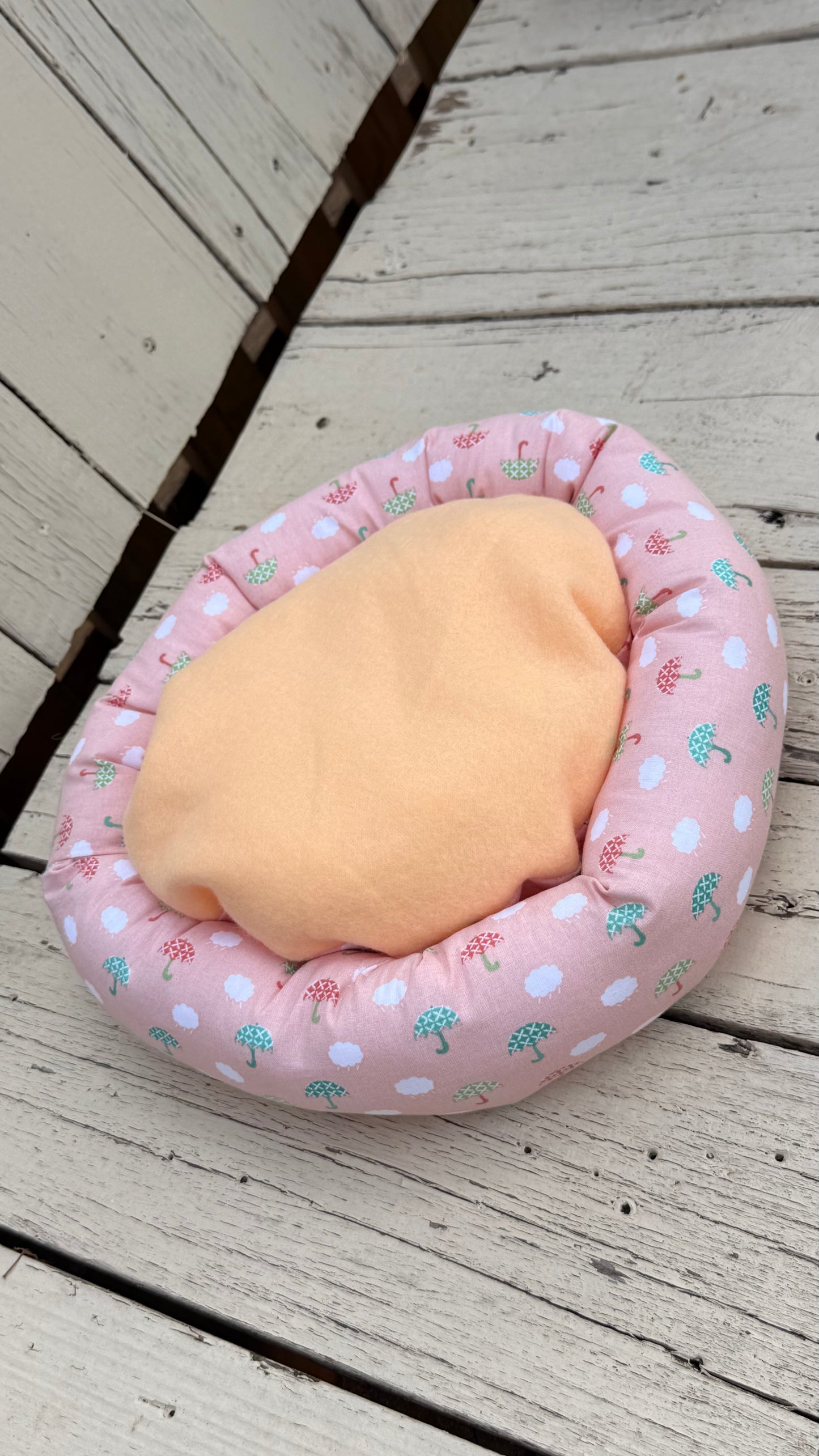 Ein runder, handgemachter Kuschel-Donut für Meerschweinchen aus altrosa Baumwollstoff. Das Material ist durchgehend mit einem Muster aus kleinen, weißen Wolken sowie grünen und roten Regenschirmen bedruckt. Das Kleintierbett liegt flach auf einer weiß gestrichenen Holzbank.