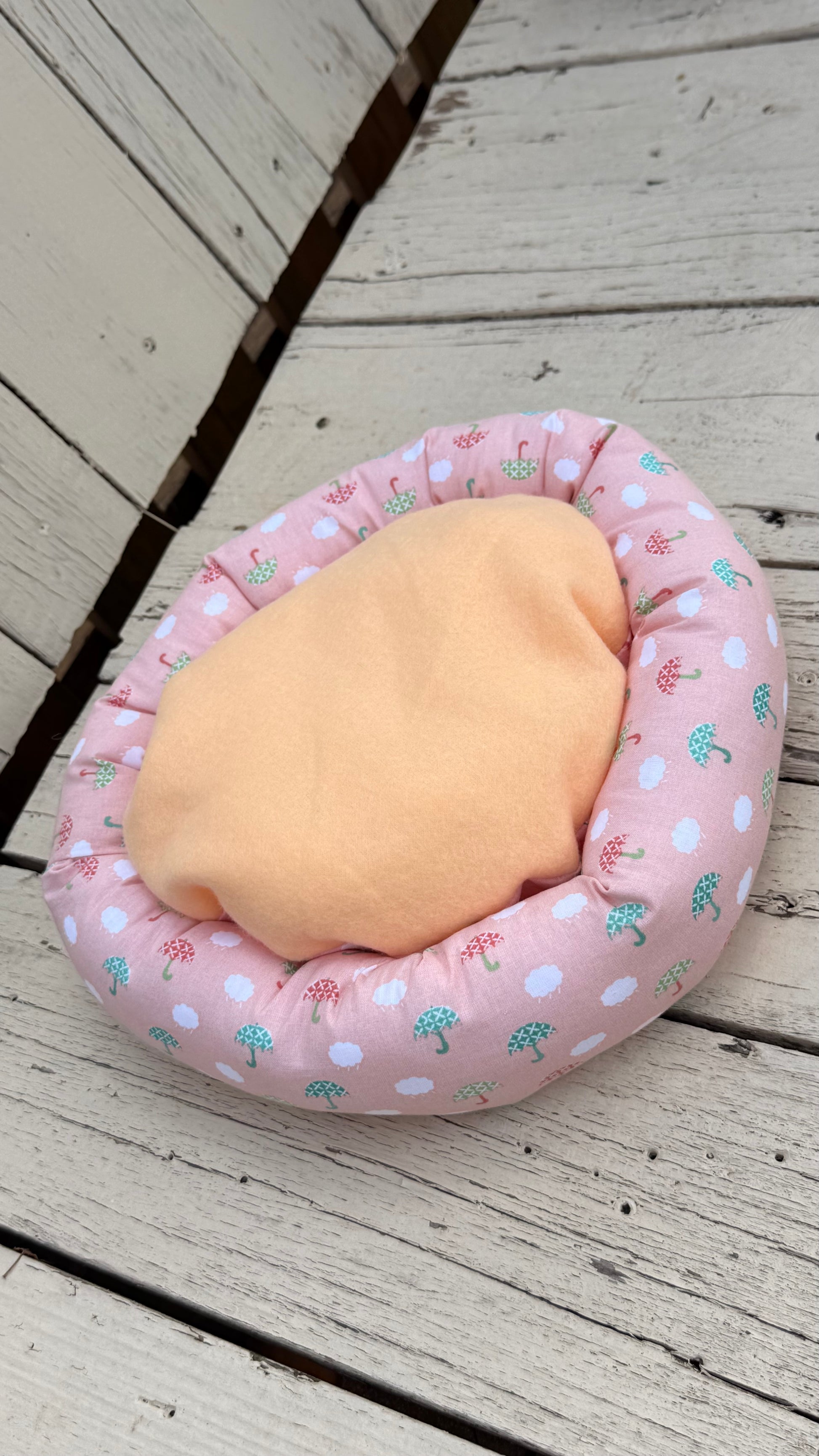 Ein runder, handgemachter Kuschel-Donut für Meerschweinchen aus altrosa Baumwollstoff. Das Material ist durchgehend mit einem Muster aus kleinen, weißen Wolken sowie grünen und roten Regenschirmen bedruckt. Das Kleintierbett liegt flach auf einer weiß gestrichenen Holzbank.