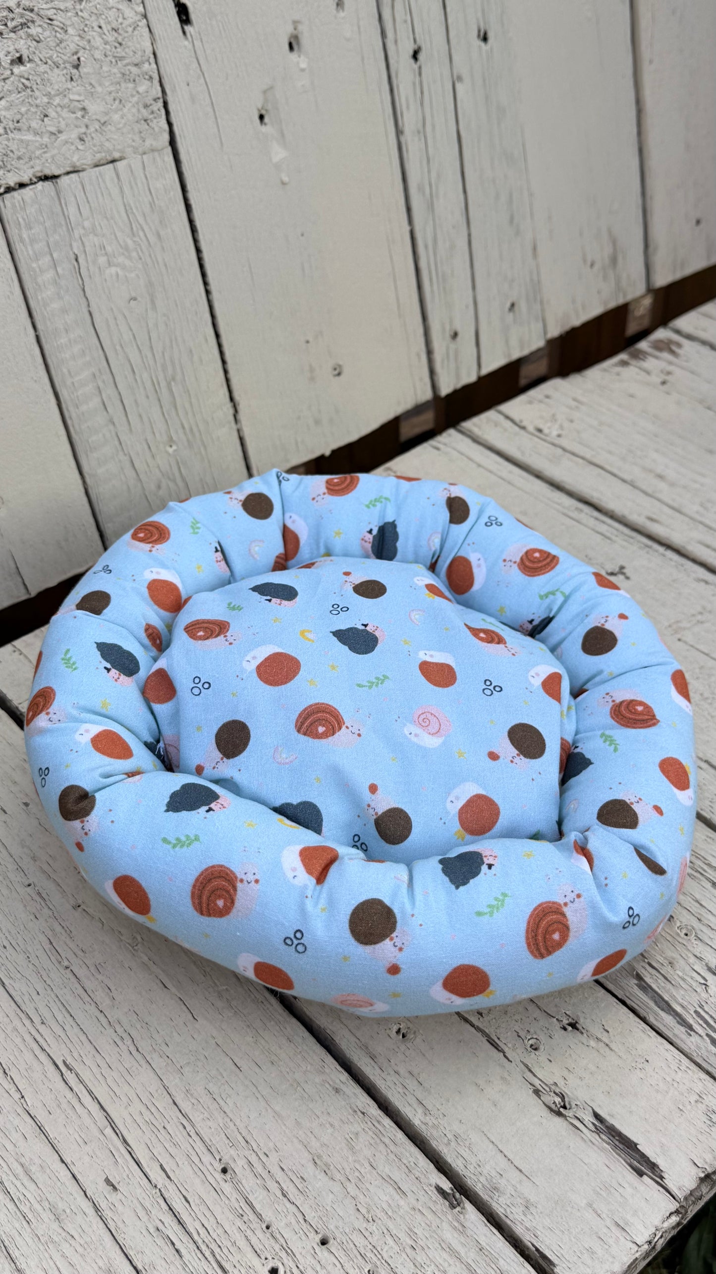 Ein runder, handgemachter Kuschel-Donut für Meerschweinchen aus hellblauem Baumwollstoff. Der Stoff ist komplett mit niedlichen Motiven von kleinen Igeln, bunten Schneckenhäusern und Pilzen bedruckt. Das gemütliche Kleintierbett liegt flach auf einer weiß gestrichenen Holzbank
