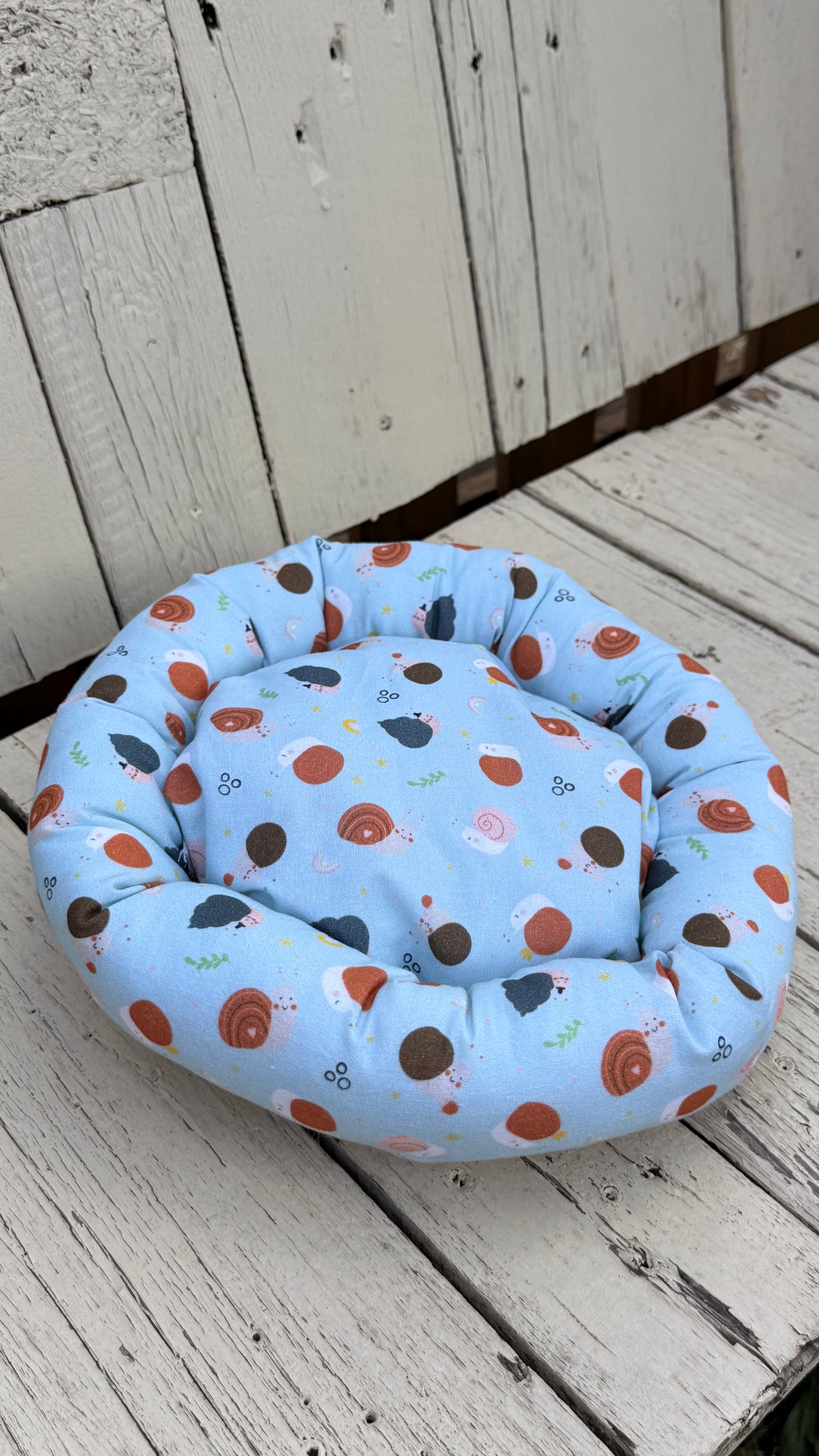 Ein runder, handgemachter Kuschel-Donut für Meerschweinchen aus hellblauem Baumwollstoff. Der Stoff ist komplett mit niedlichen Motiven von kleinen Igeln, bunten Schneckenhäusern und Pilzen bedruckt. Das gemütliche Kleintierbett liegt flach auf einer weiß gestrichenen Holzbank