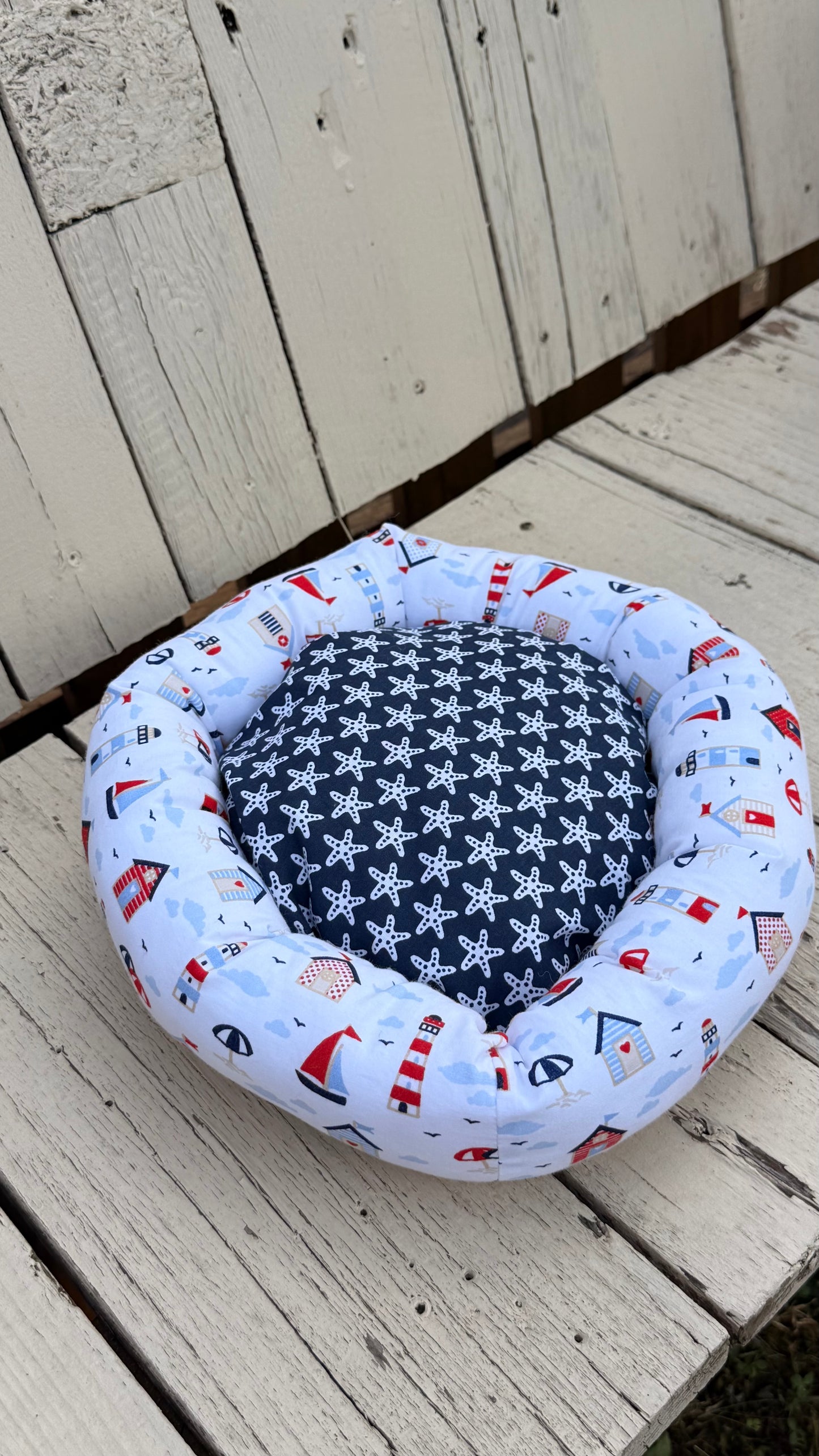 Ein handgemachter, runder Kuschel-Donut für Meerschweinchen im maritimen Design. Der äußere Ring besteht aus weißem Baumwollstoff, bedruckt mit bunten Leuchttürmen, Segelbooten, Strandhäusern und Sonnenschirmen. Die weiche Liegemulde im Inneren ist mit einem blau-grauen Stoff mit weißen Seesternen gemustert. Das Kleintierbett liegt auf einer rustikalen, weißen Holzbank