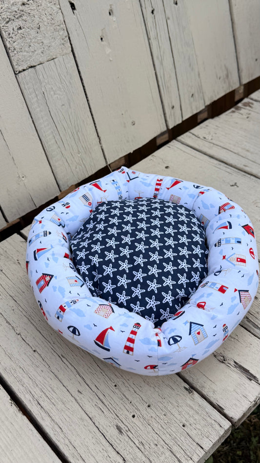 Ein handgemachter, runder Kuschel-Donut für Meerschweinchen im maritimen Design. Der äußere Ring besteht aus weißem Baumwollstoff, bedruckt mit bunten Leuchttürmen, Segelbooten, Strandhäusern und Sonnenschirmen. Die weiche Liegemulde im Inneren ist mit einem blau-grauen Stoff mit weißen Seesternen gemustert. Das Kleintierbett liegt auf einer rustikalen, weißen Holzbank