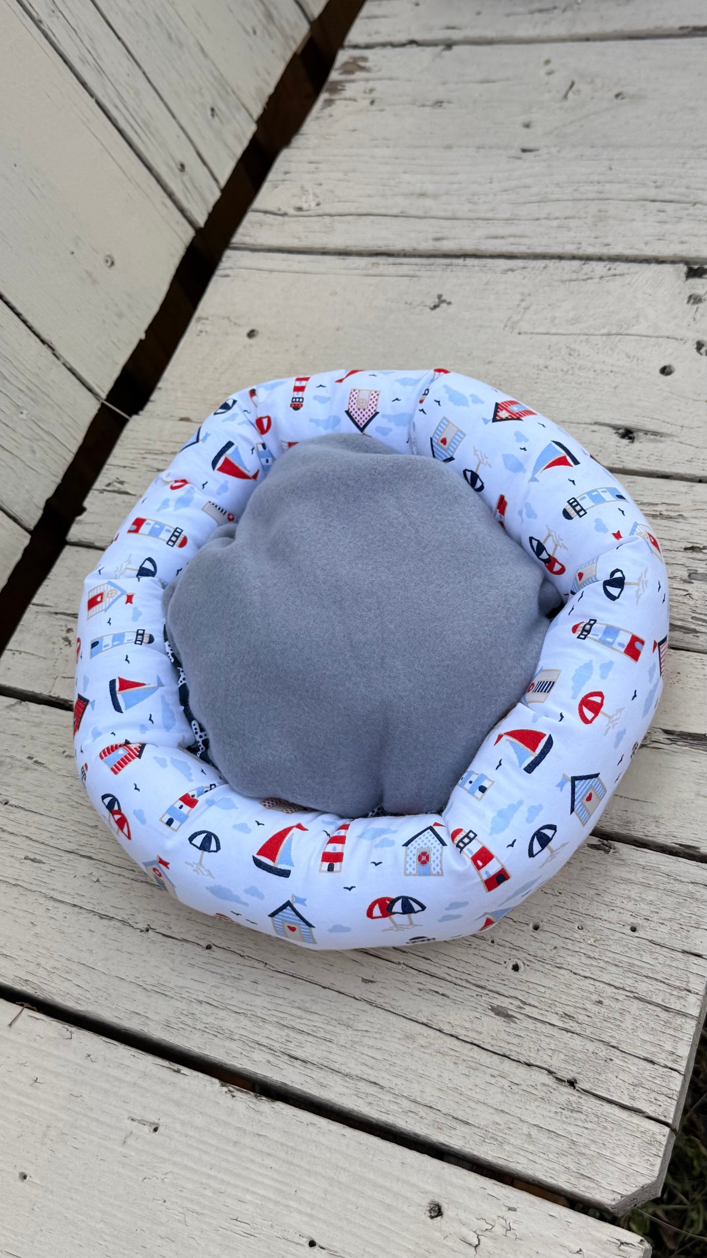 Ein handgemachter, runder Kuschel-Donut für Meerschweinchen im maritimen Design. Der äußere Ring besteht aus weißem Baumwollstoff, bedruckt mit bunten Leuchttürmen, Segelbooten, Strandhäusern und Sonnenschirmen. Die weiche Liegemulde im Inneren ist mit einem blau-grauen Stoff mit weißen Seesternen gemustert. Das Kleintierbett liegt auf einer rustikalen, weißen Holzbank