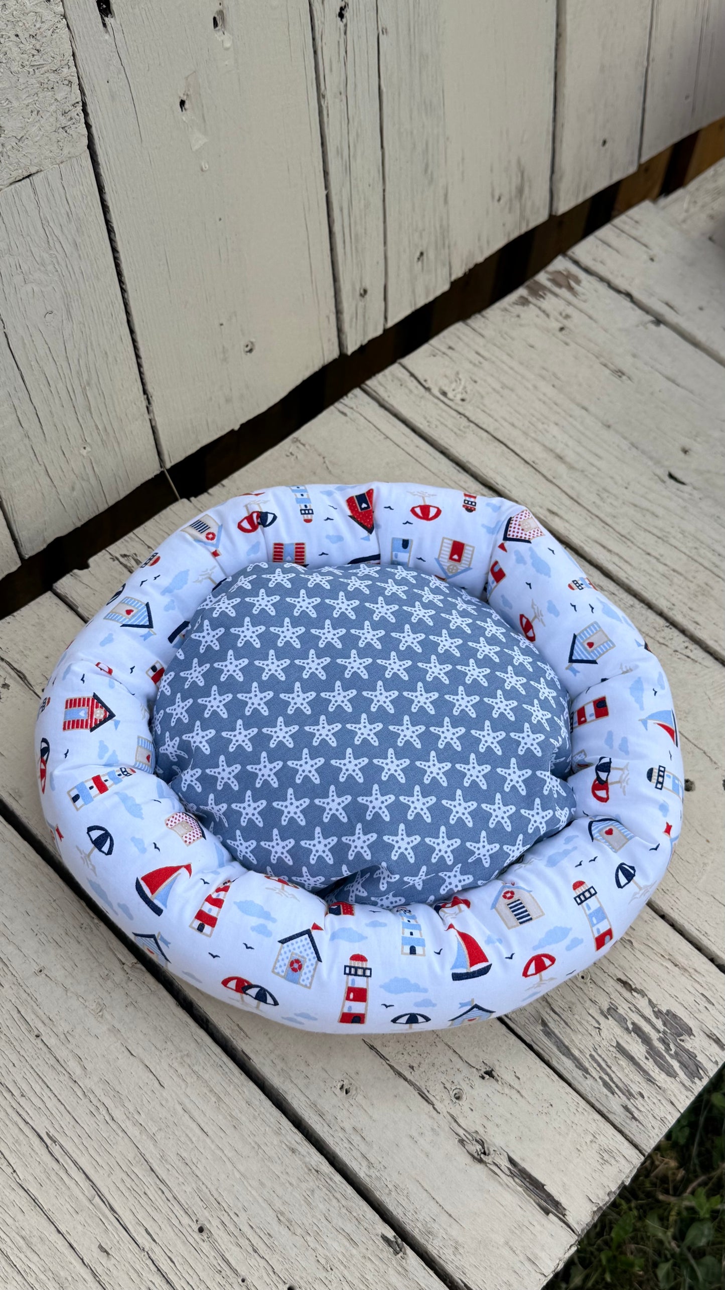 Ein handgemachter, runder Kuschel-Donut für Meerschweinchen im maritimen Design. Der äußere Ring besteht aus weißem Baumwollstoff, bedruckt mit bunten Leuchttürmen, Segelbooten, Strandhäusern und Sonnenschirmen. Die weiche Liegemulde im Inneren ist mit einem blau-grauen Stoff mit weißen Seesternen gemustert. Das Kleintierbett liegt auf einer rustikalen, weißen Holzbank