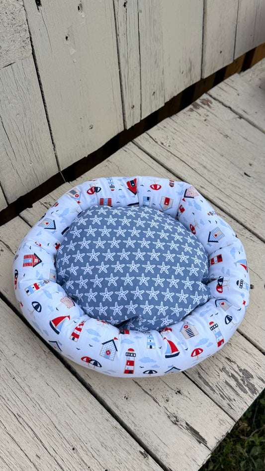 Ein handgemachter, runder Kuschel-Donut für Meerschweinchen im maritimen Design. Der äußere Ring besteht aus weißem Baumwollstoff, bedruckt mit bunten Leuchttürmen, Segelbooten, Strandhäusern und Sonnenschirmen. Die weiche Liegemulde im Inneren ist mit einem blau-grauen Stoff mit weißen Seesternen gemustert. Das Kleintierbett liegt auf einer rustikalen, weißen Holzbank