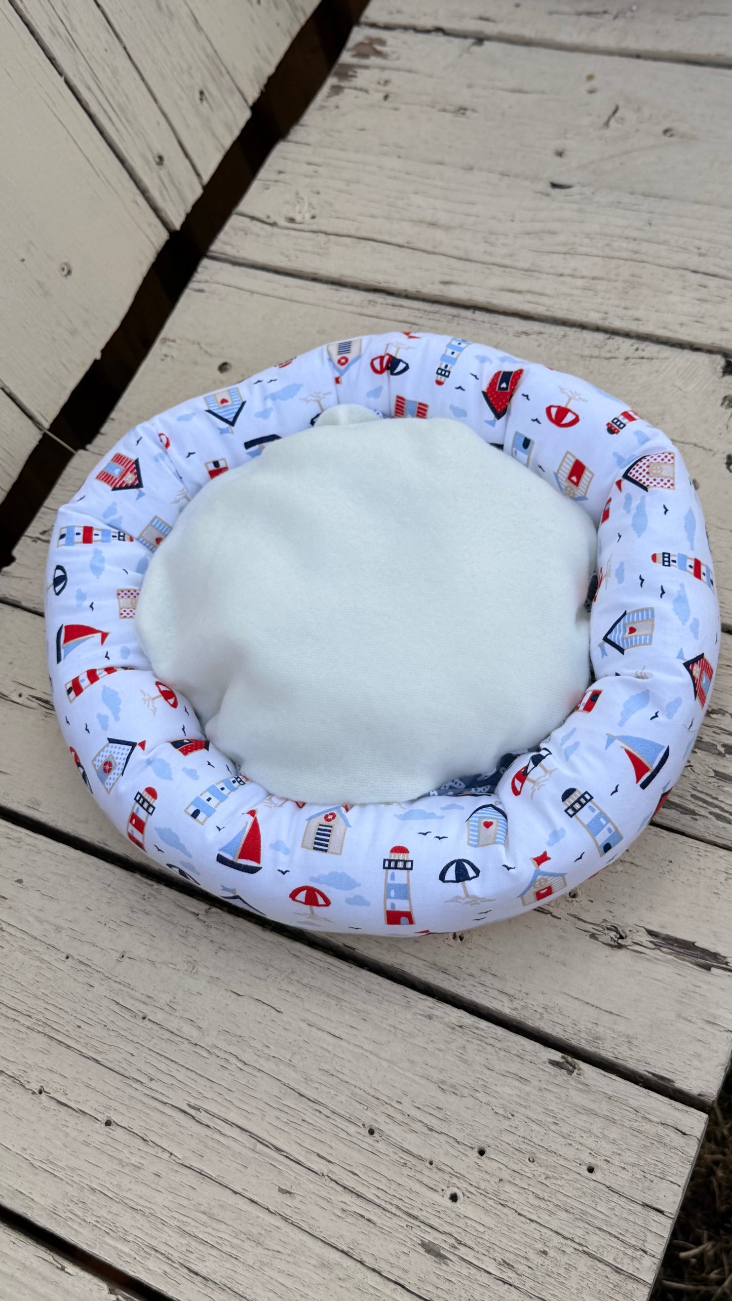 Ein handgemachter, runder Kuschel-Donut für Meerschweinchen im maritimen Design. Der äußere Ring besteht aus weißem Baumwollstoff, bedruckt mit bunten Leuchttürmen, Segelbooten, Strandhäusern und Sonnenschirmen. Die weiche Liegemulde im Inneren ist mit einem blau-grauen Stoff mit weißen Seesternen gemustert. Das Kleintierbett liegt auf einer rustikalen, weißen Holzbank