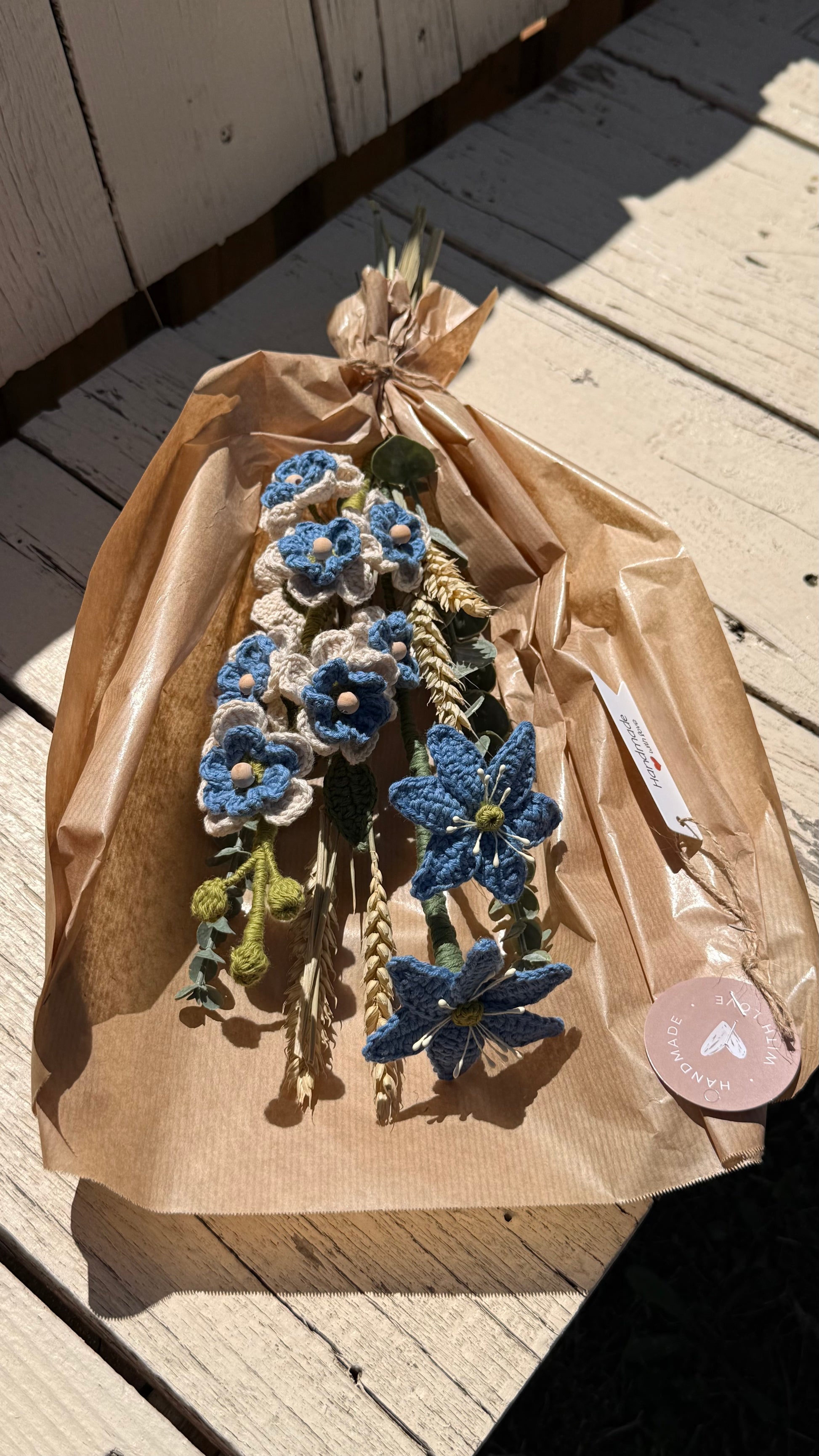 Handgefertigter Blumenstrauß aus der Mama Edeltrud Kollektion mit gehäkelten blauen und cremefarbenen Blüten sowie Weizenähren, liebevoll in braunem Papier verpackt als nachhaltige Deko-Blumen