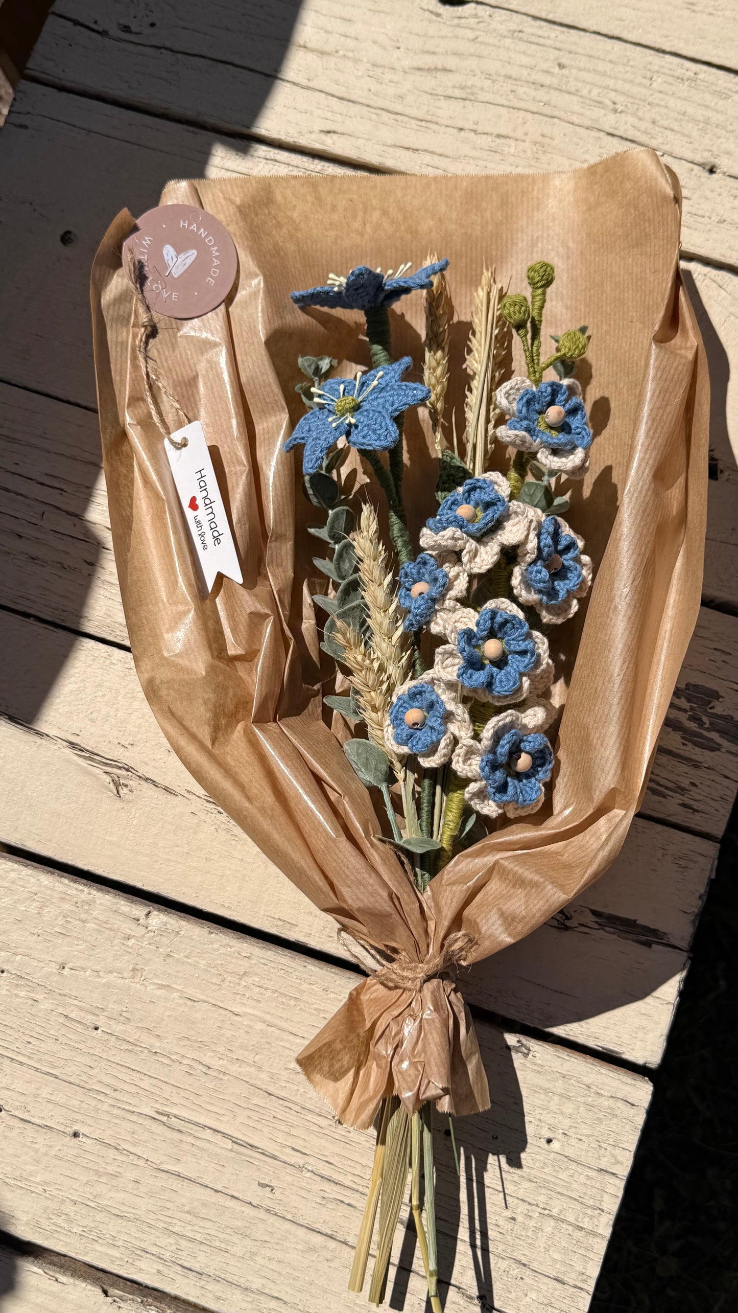 Handgefertigter Blumenstrauß aus der Mama Edeltrud Kollektion mit gehäkelten blauen und cremefarbenen Blüten sowie Weizenähren, liebevoll in braunem Papier verpackt als nachhaltige Deko-Blumen