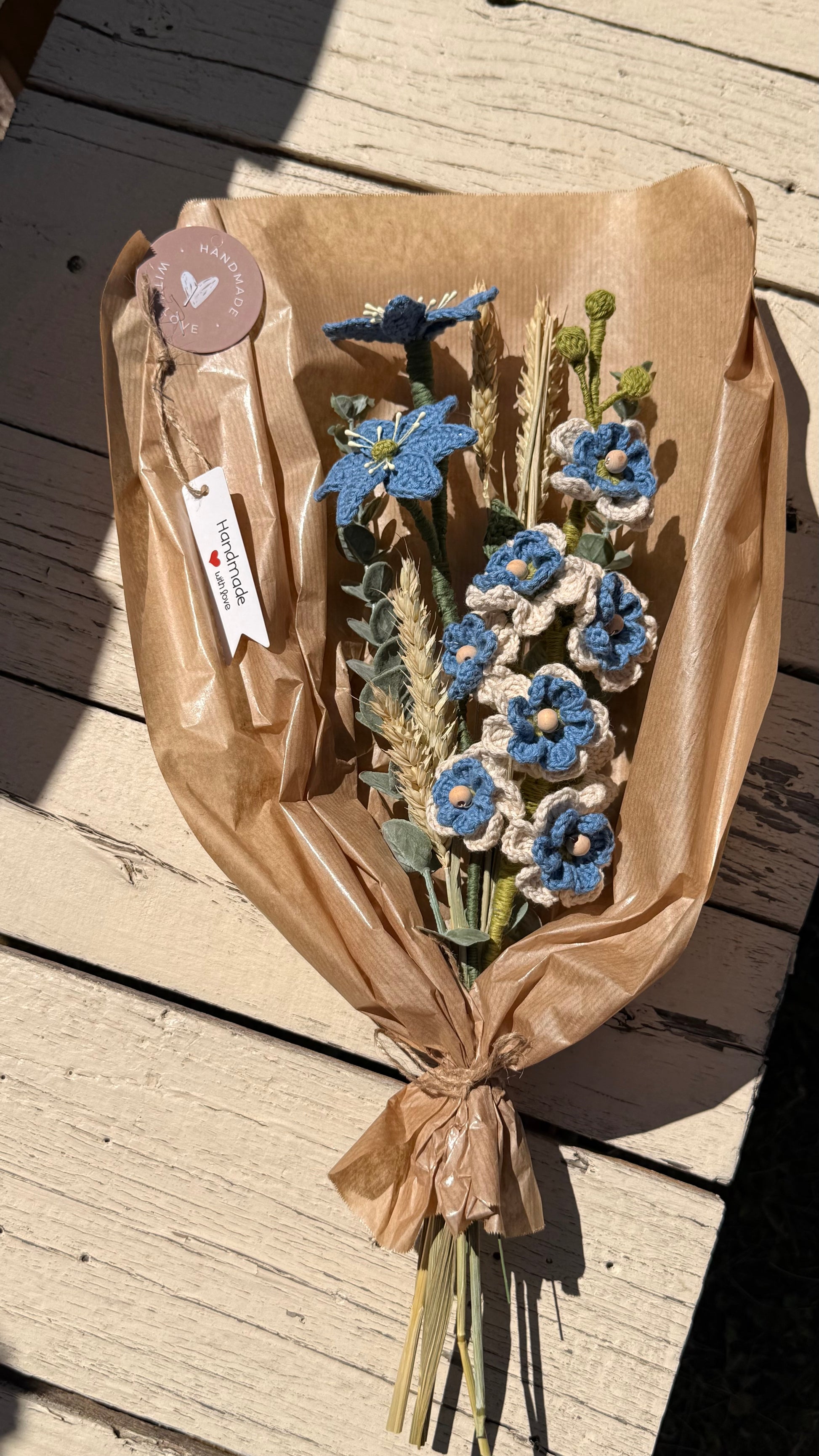 Handgefertigter Blumenstrauß aus der Mama Edeltrud Kollektion mit gehäkelten blauen und cremefarbenen Blüten sowie Weizenähren, liebevoll in braunem Papier verpackt als nachhaltige Deko-Blumen
