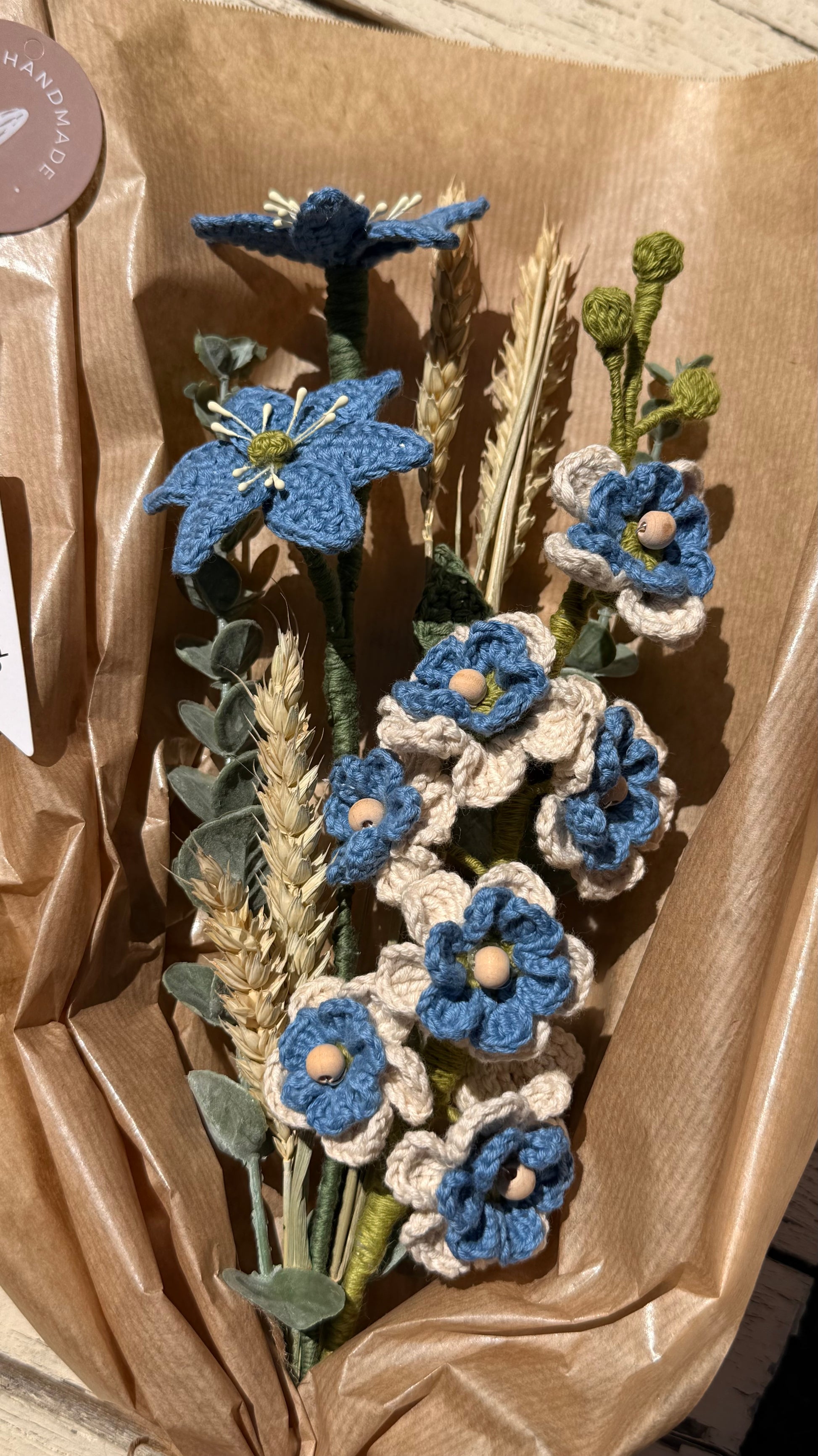 Handgefertigter Blumenstrauß aus der Mama Edeltrud Kollektion mit gehäkelten blauen und cremefarbenen Blüten sowie Weizenähren, liebevoll in braunem Papier verpackt als nachhaltige Deko-Blumen