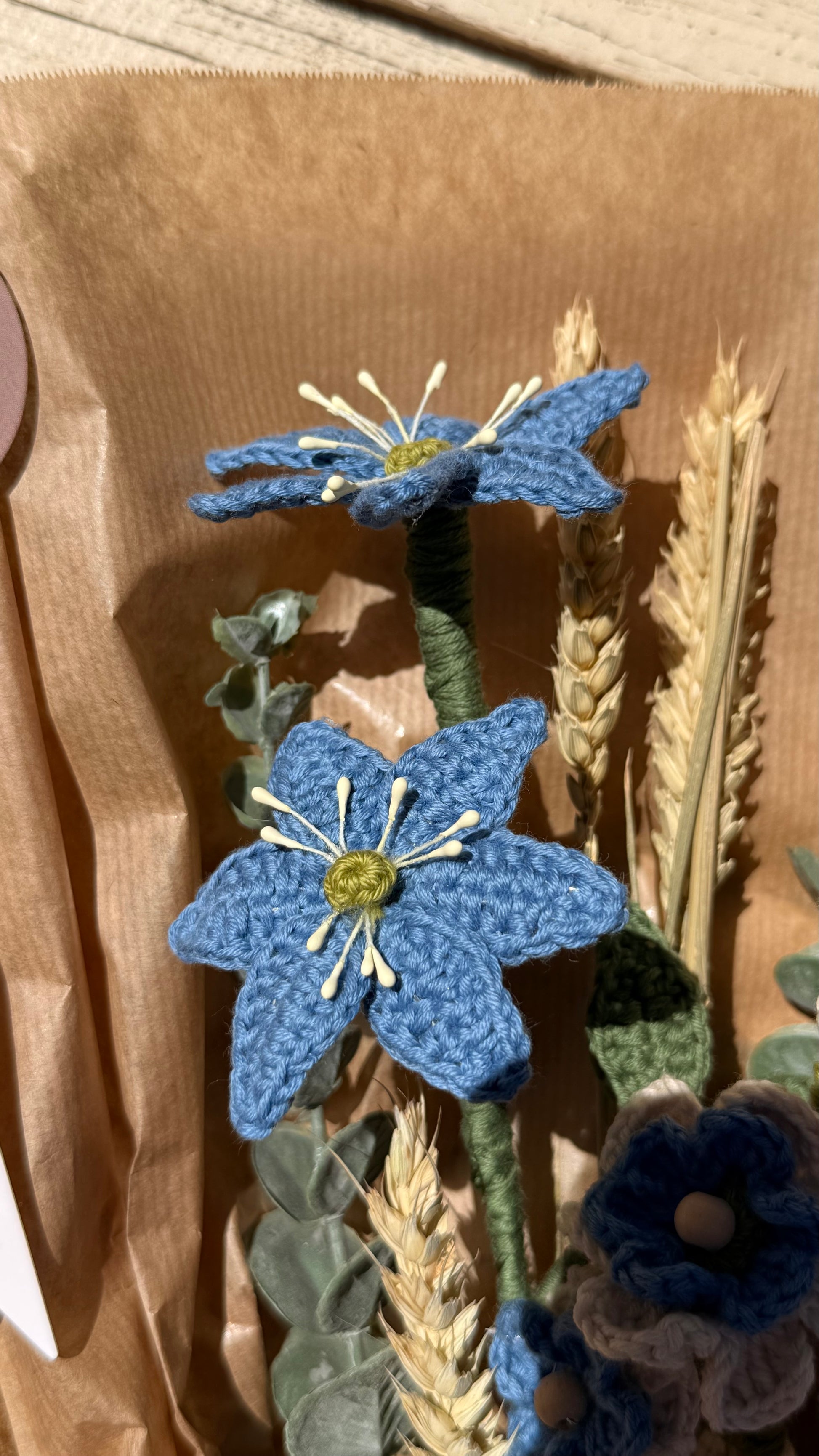 Handgefertigter Blumenstrauß aus der Mama Edeltrud Kollektion mit gehäkelten blauen und cremefarbenen Blüten sowie Weizenähren, liebevoll in braunem Papier verpackt als nachhaltige Deko-Blumen