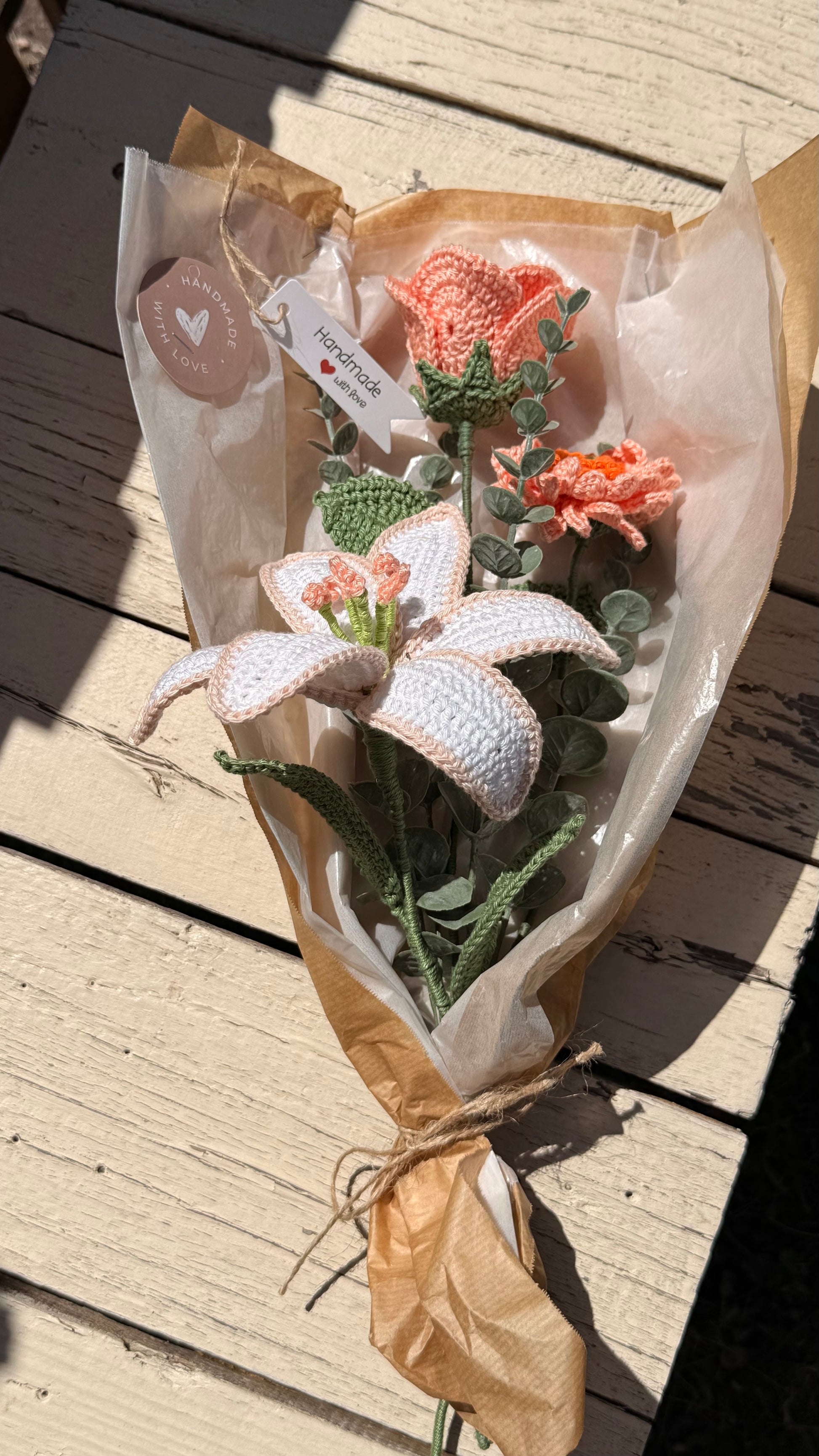 Handgefertigter Blumenstrauß aus der Mama Edeltrud Kollektion mit gehäkelter Lilie und Rosen in Pfirsich-Tönen, liebevoll verpackt in Naturpapier als nachhaltiges Geschenk.