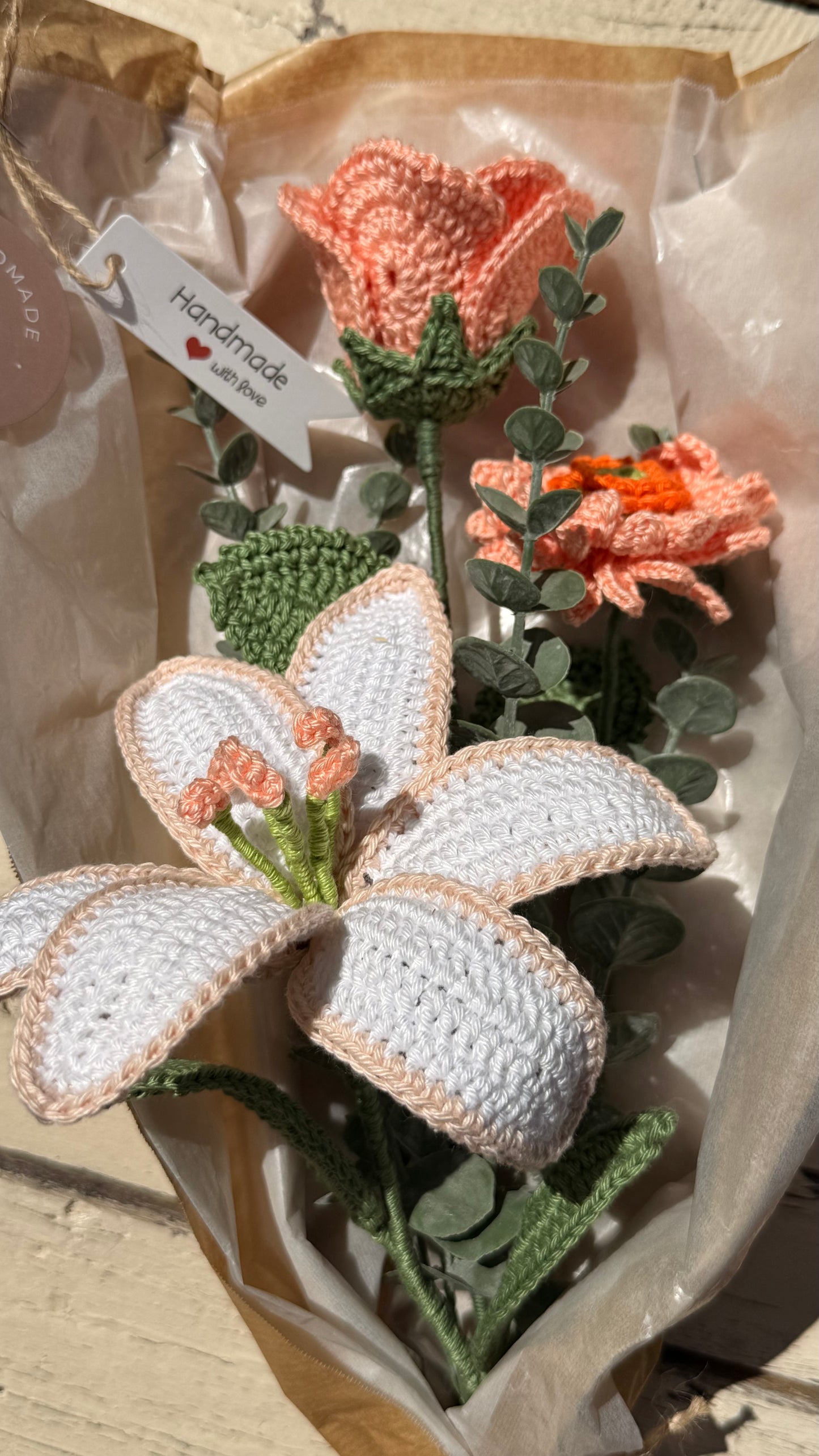 Handgefertigter Blumenstrauß aus der Mama Edeltrud Kollektion mit gehäkelter Lilie und Rosen in Pfirsich-Tönen, liebevoll verpackt in Naturpapier als nachhaltiges Geschenk.