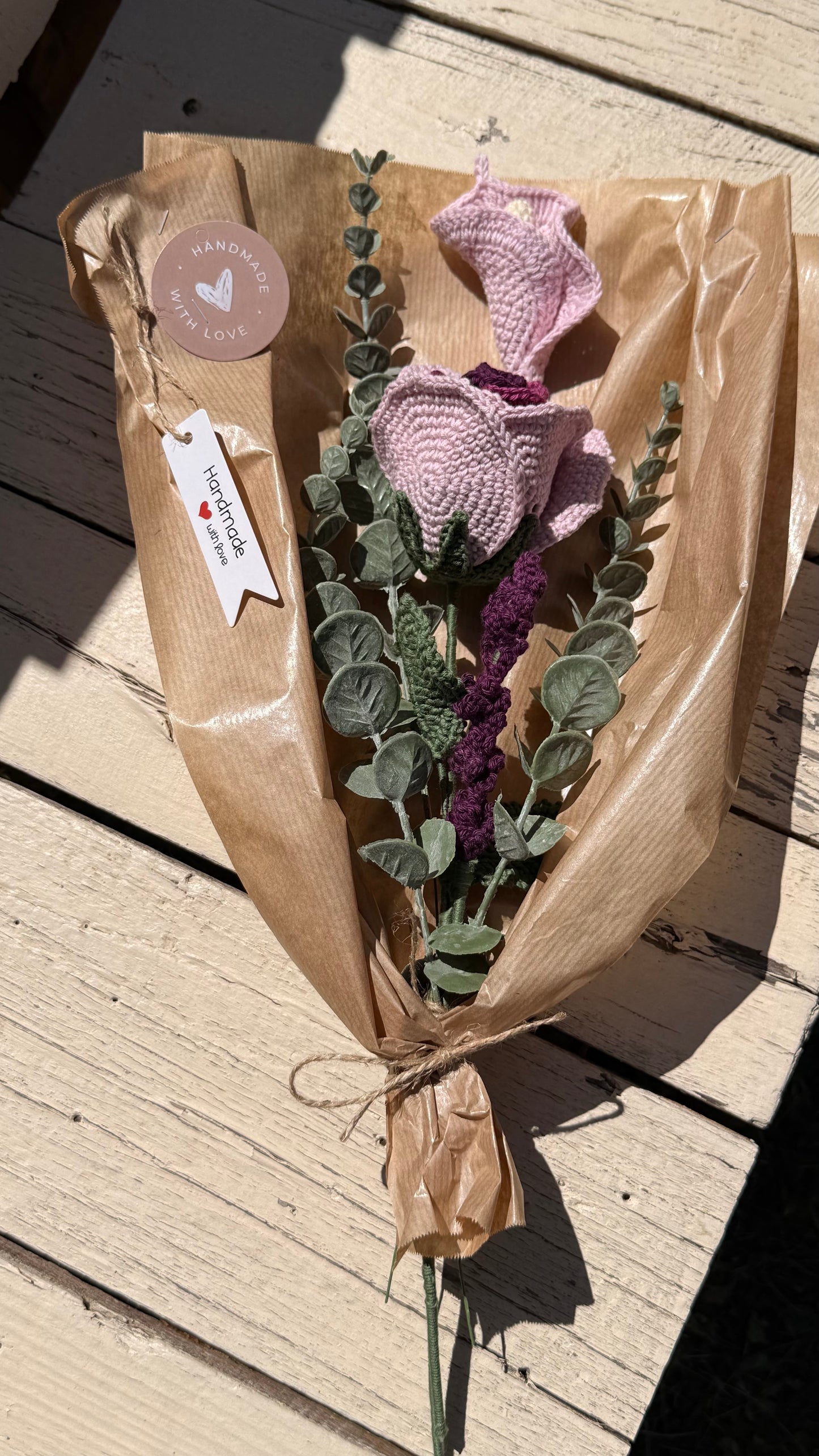 Handgefertigter Blumenstrauß aus der Mama Edeltrud Kollektion mit gehäkelten Rosen und Blüten in Rosé- und Violetttönen, elegant verpackt in Naturpapier als nachhaltiges Deko-Geschenk.
