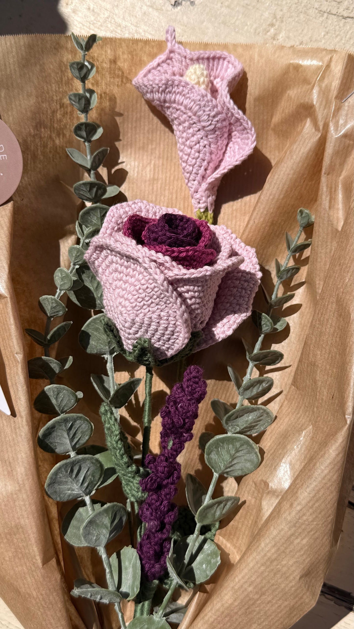 Handgefertigter Blumenstrauß aus der Mama Edeltrud Kollektion mit gehäkelten Rosen und Blüten in Rosé- und Violetttönen, elegant verpackt in Naturpapier als nachhaltiges Deko-Geschenk.