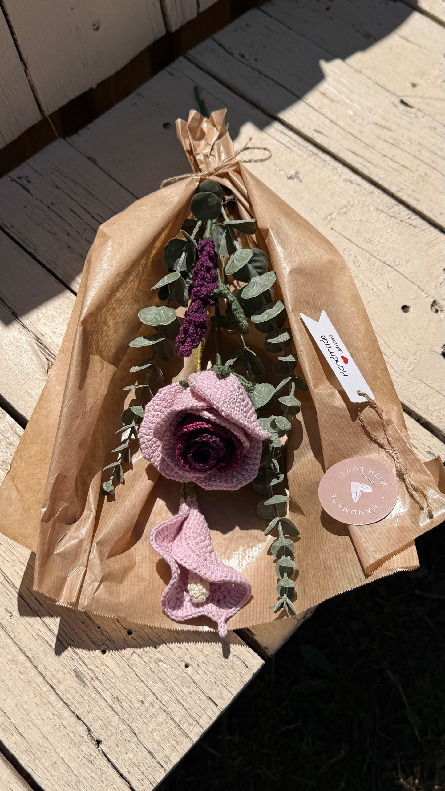 Handgefertigter Blumenstrauß aus der Mama Edeltrud Kollektion mit gehäkelten Rosen und Blüten in Rosé- und Violetttönen, elegant verpackt in Naturpapier als nachhaltiges Deko-Geschenk.